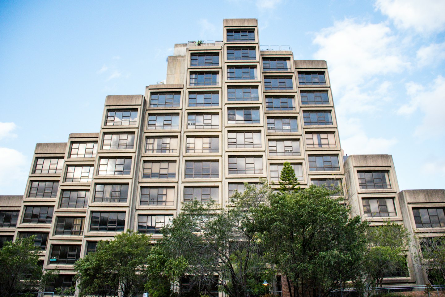 Stepped concrete building with rectangular windows behind green trees.